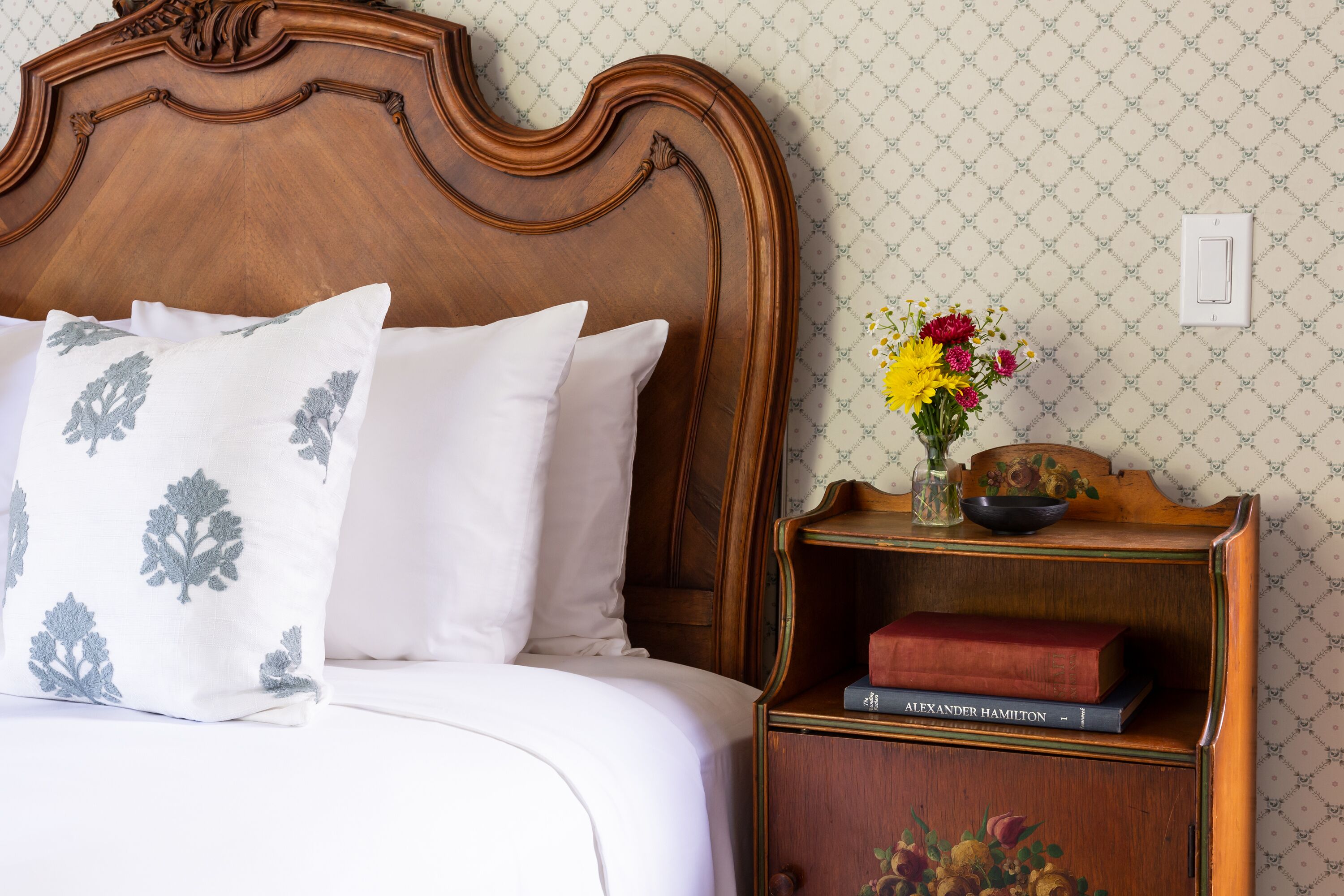 Een keurig opgemaakt bed met witte beddengoed en decoratieve kussens, een houten nachtkastje met een bloevas en boeken, en een behang met patroon.