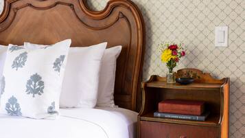 Een keurig opgemaakt bed met witte beddengoed en decoratieve kussens, een houten nachtkastje met een bloevas en boeken, en een behang met patroon.