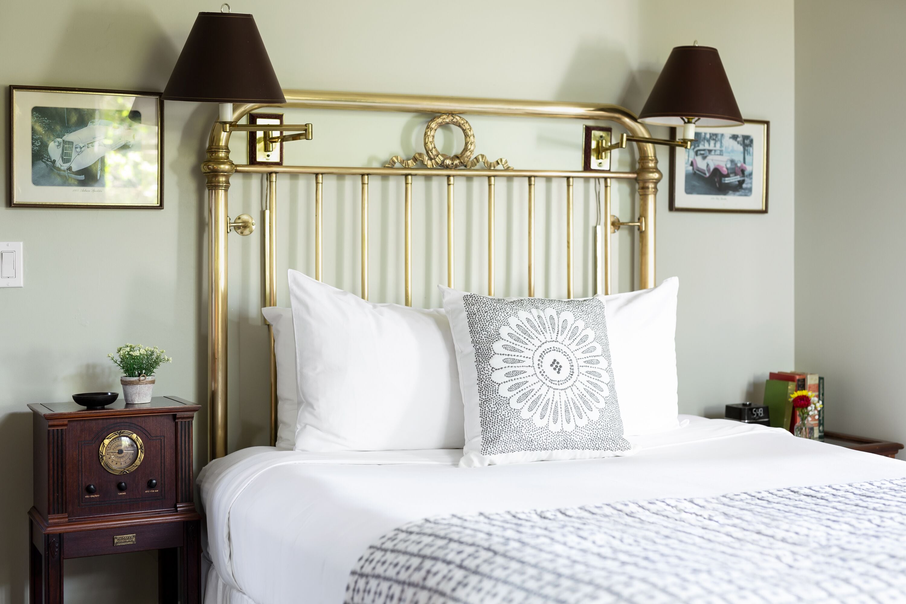 A neatly made bed with white linens and two pillows, a small wooden nightstand with a radio, and a wall-mounted lamp.