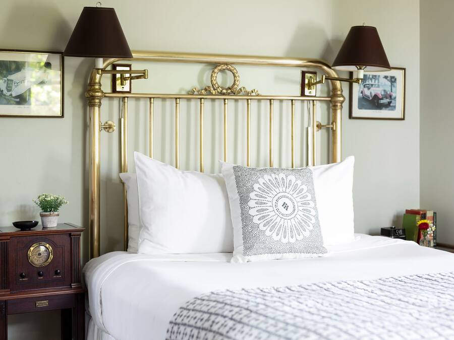 A neatly made bed with white linens and two pillows, a small wooden nightstand with a radio, and a wall-mounted lamp.