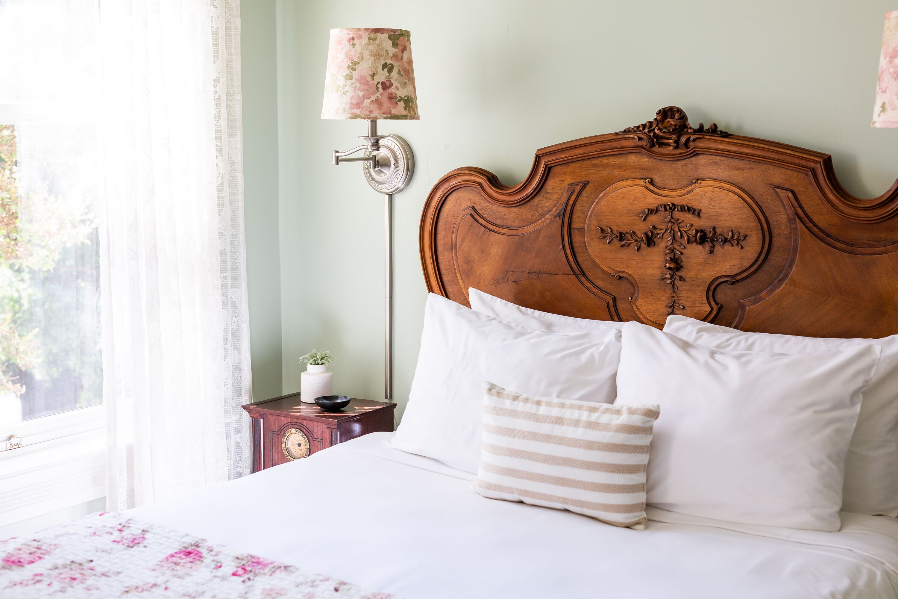 A neatly made bed with white linens and striped pillows, a wooden headboard with intricate carvings, a bedside table with a lamp, and a window with sheer curtains.