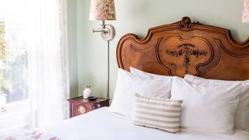A neatly made bed with white linens and striped pillows, a wooden headboard with intricate carvings, a bedside table with a lamp, and a window with sheer curtains.