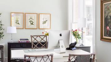 Een kamer met een wit bureau, twee rieten stoelen, een laptop en ingelijste afbeeldingen van planten aan de muur.