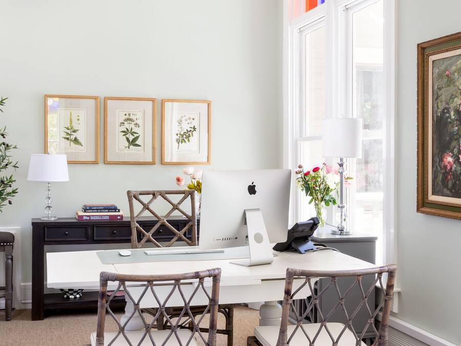 Een kamer met een wit bureau, twee rieten stoelen, een laptop en ingelijste afbeeldingen van planten aan de muur.