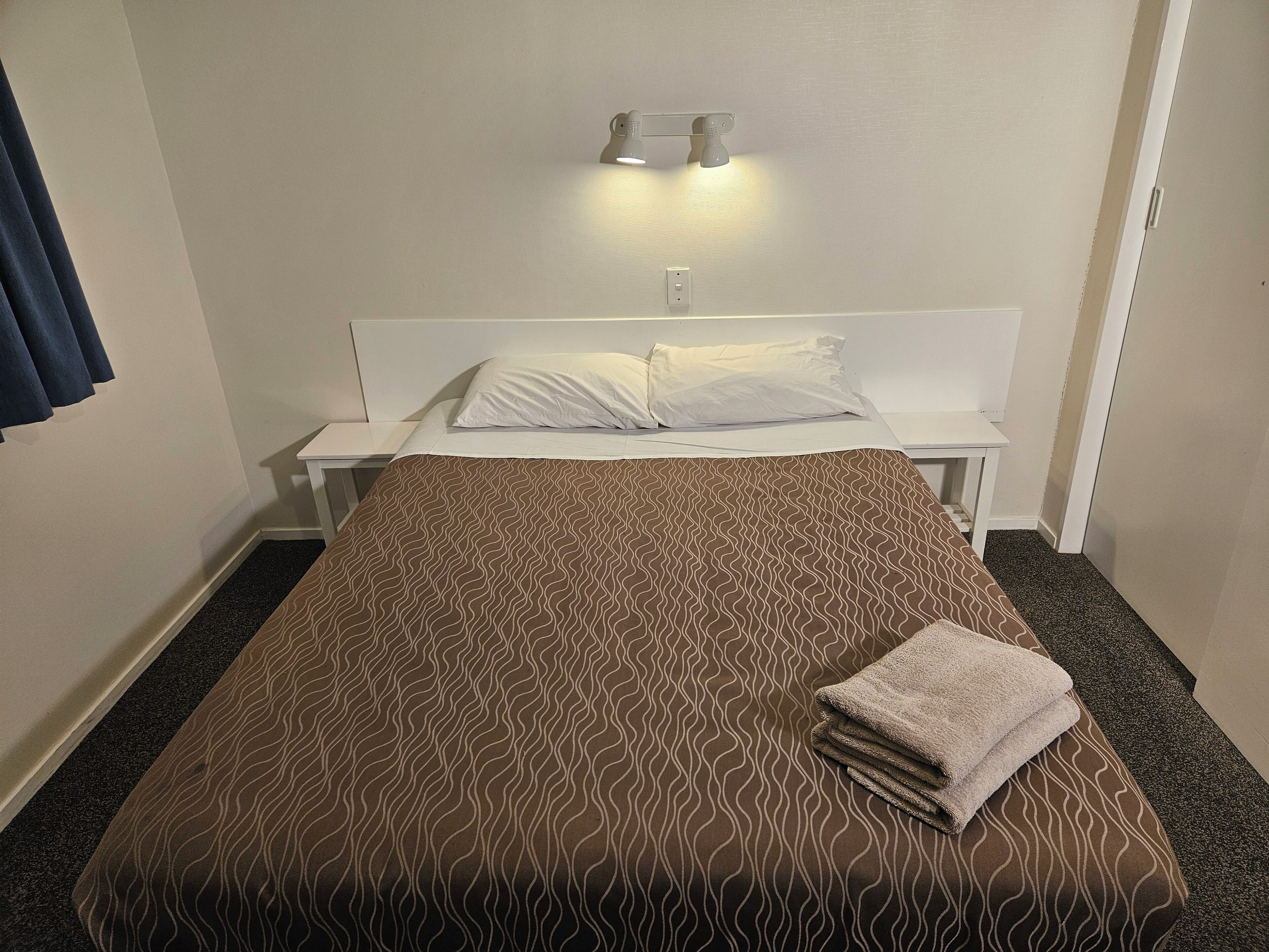 A neatly made bed with a patterned bedspread, a white pillow, and a folded towel on the side. There is a bedside table with a lamp and a wall-mounted light fixture above the bed.