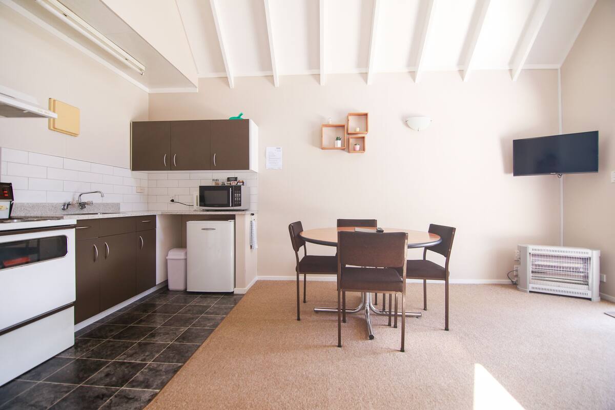 A modern kitchen with dark cabinets, a white refrigerator, and a microwave. There is a dining table with four chairs and a small plant on the wall.
