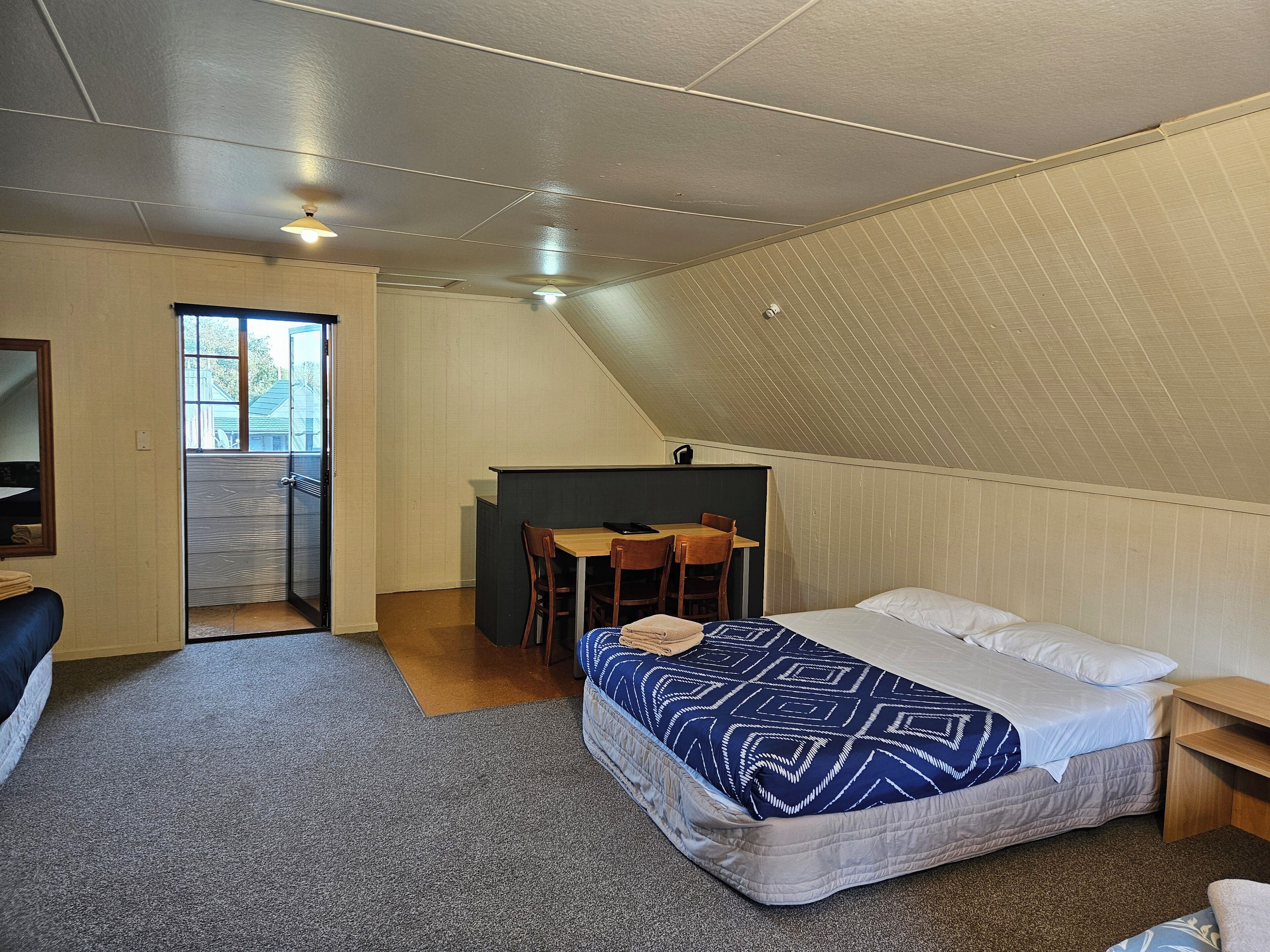 A small, cozy room with a bed, a desk, and a chair. There is a window and a door leading to another room.