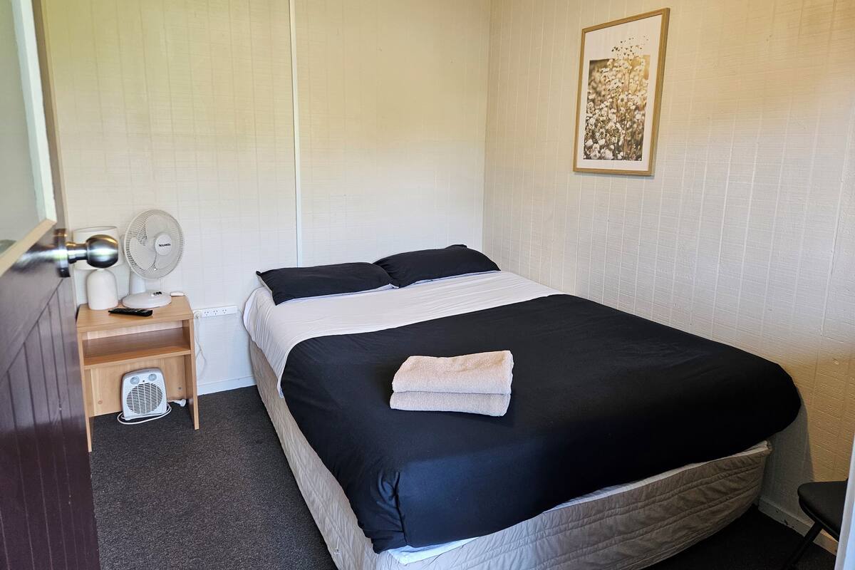 A neatly made bed with a black and white duvet, a bedside table with a fan, a remote control, and a small heater.