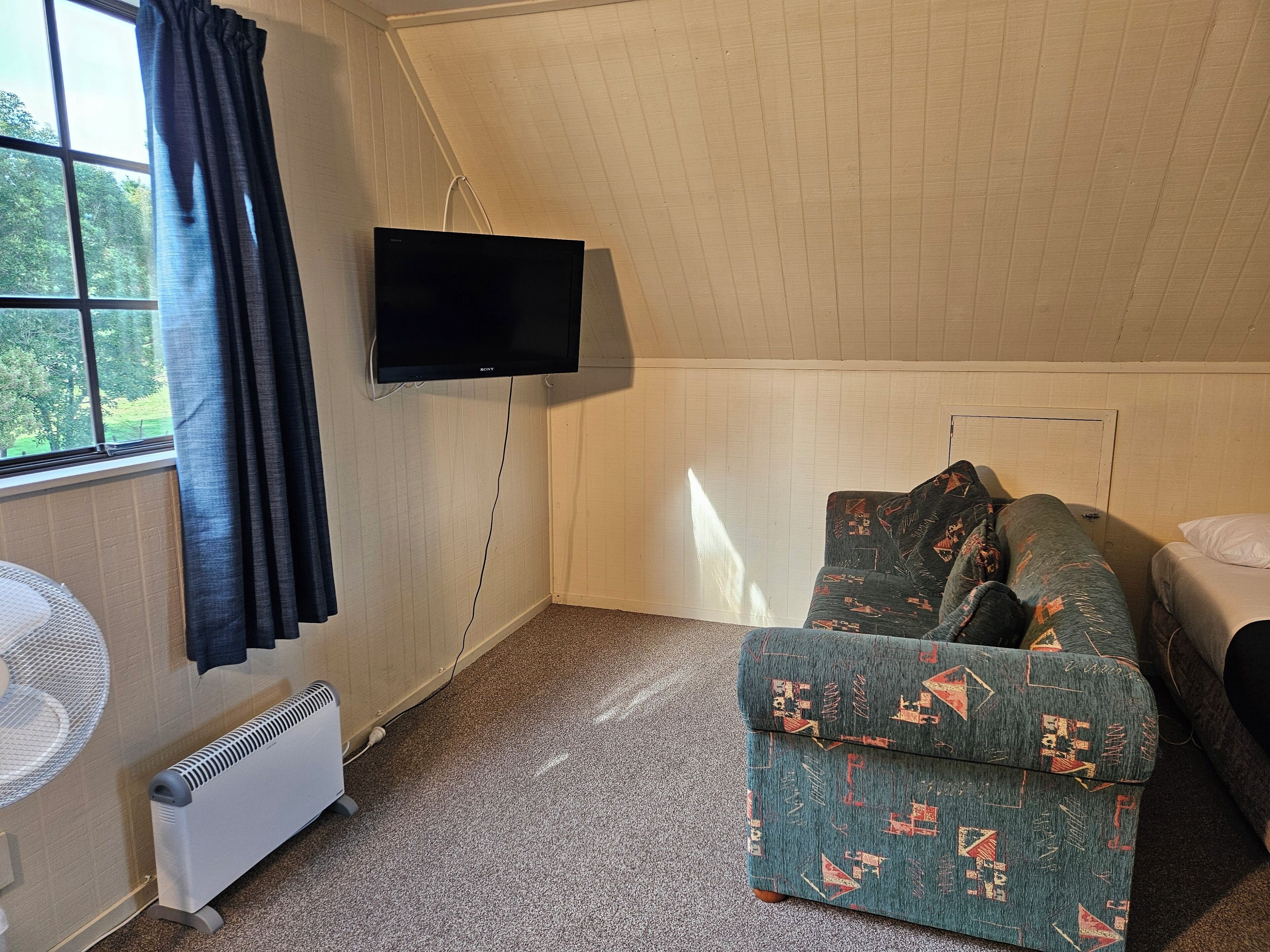 A room with a green sofa, a bed, a flat-screen TV mounted on the wall, a standing fan, and a window with curtains.