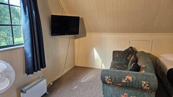 A room with a green sofa, a bed, a flat-screen TV mounted on the wall, a standing fan, and a window with curtains.