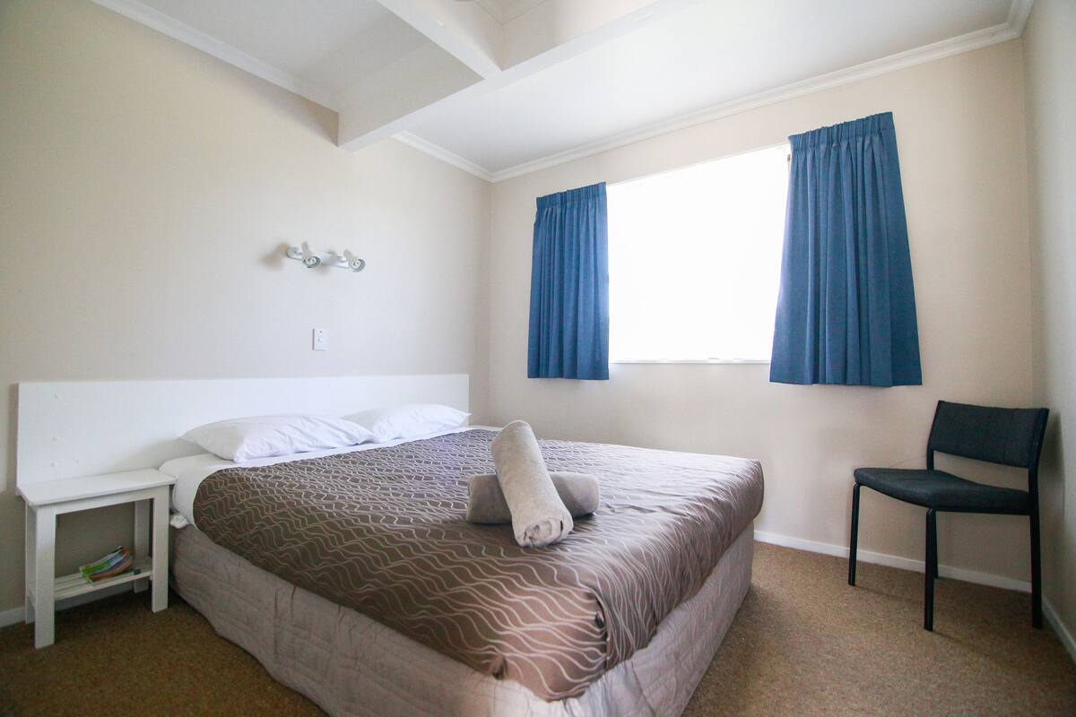 A hotel room with a bed, a chair, a nightstand, and a window with curtains.