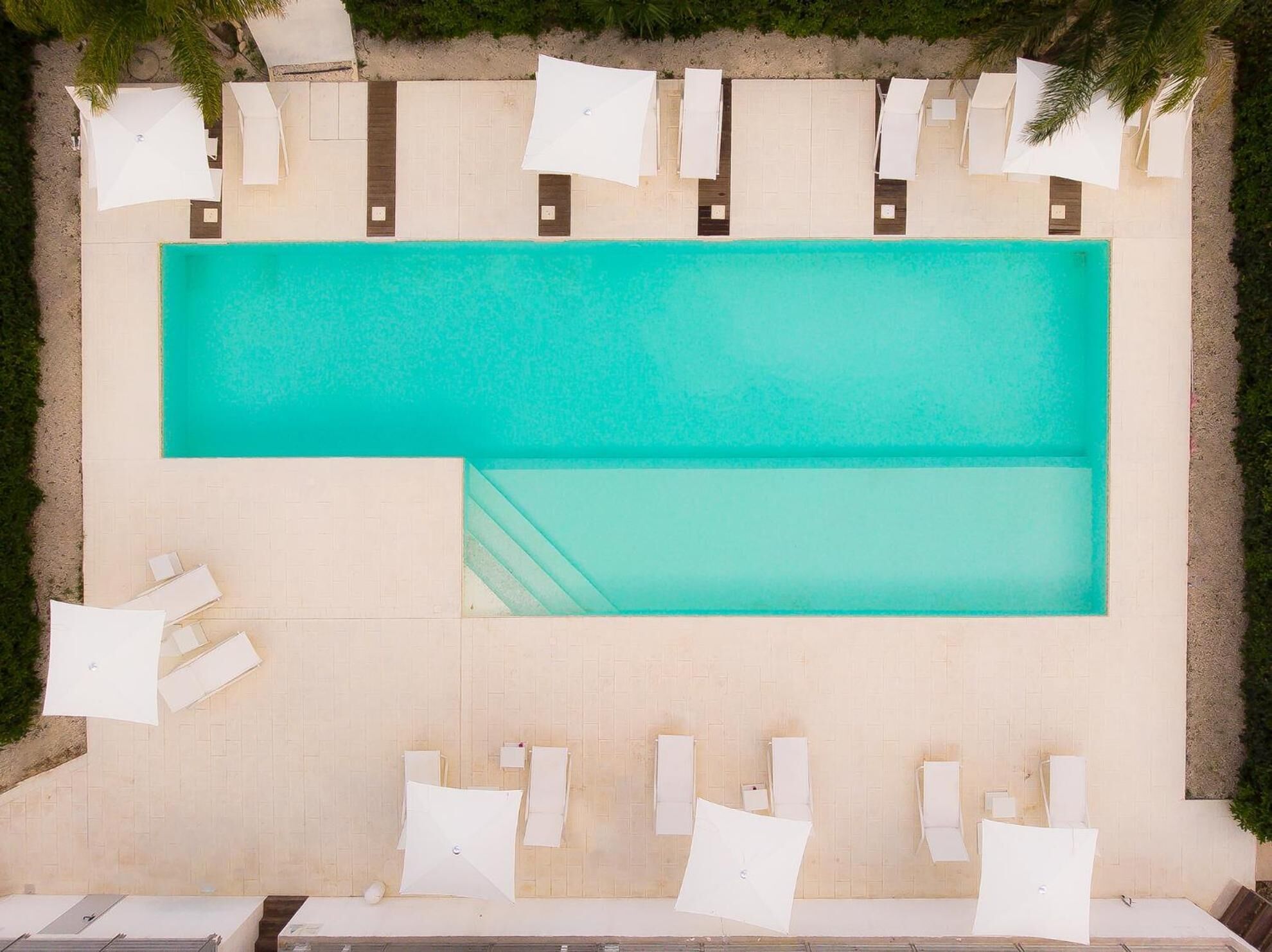 Vista aérea de uma piscina retangular com um deck revestido ao redor. Há cadeiras de lounge brancas e guarda-sóis dispostos junto à piscina.