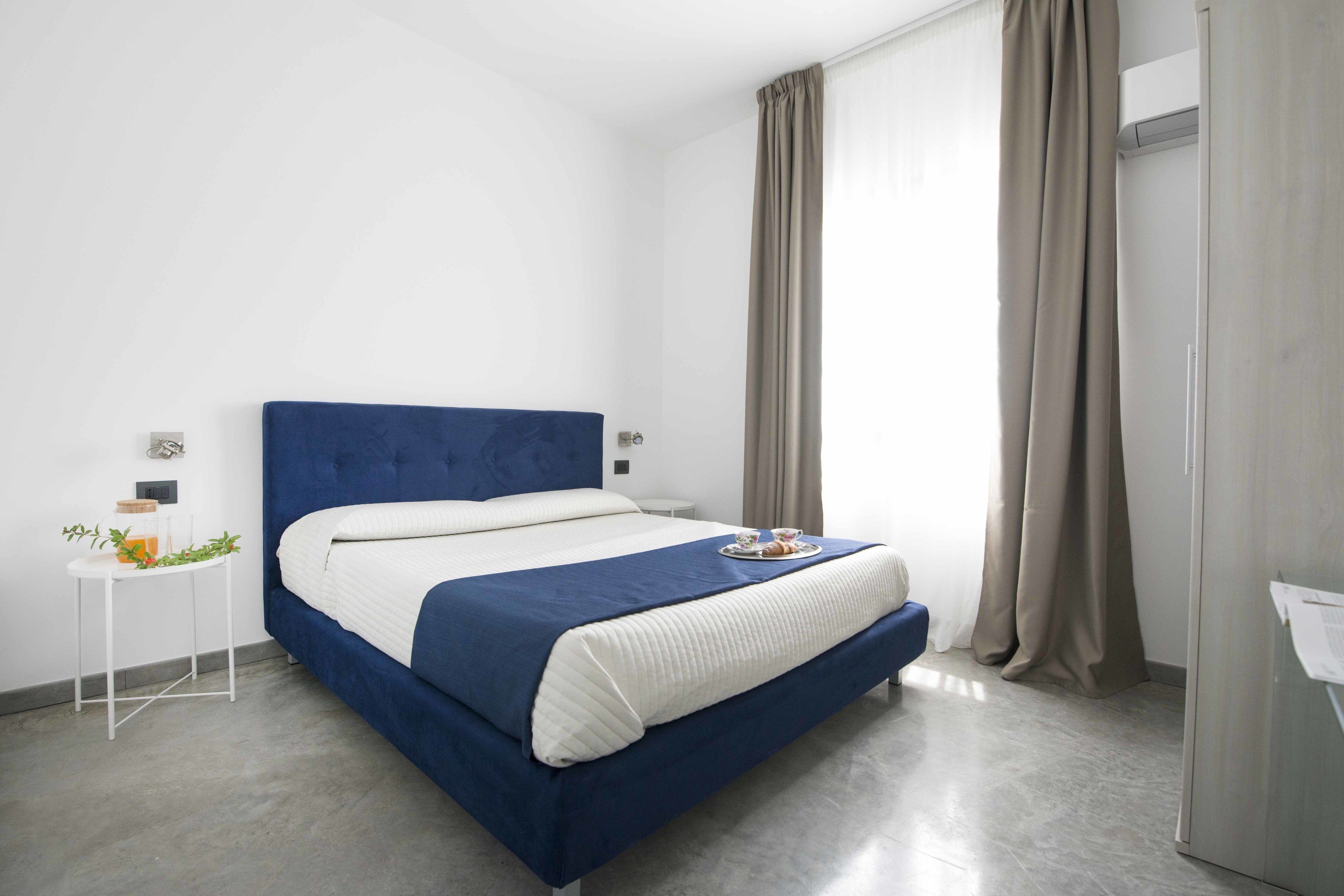 A modern hotel room with a blue bed, white walls, and a small side table with a plant.
