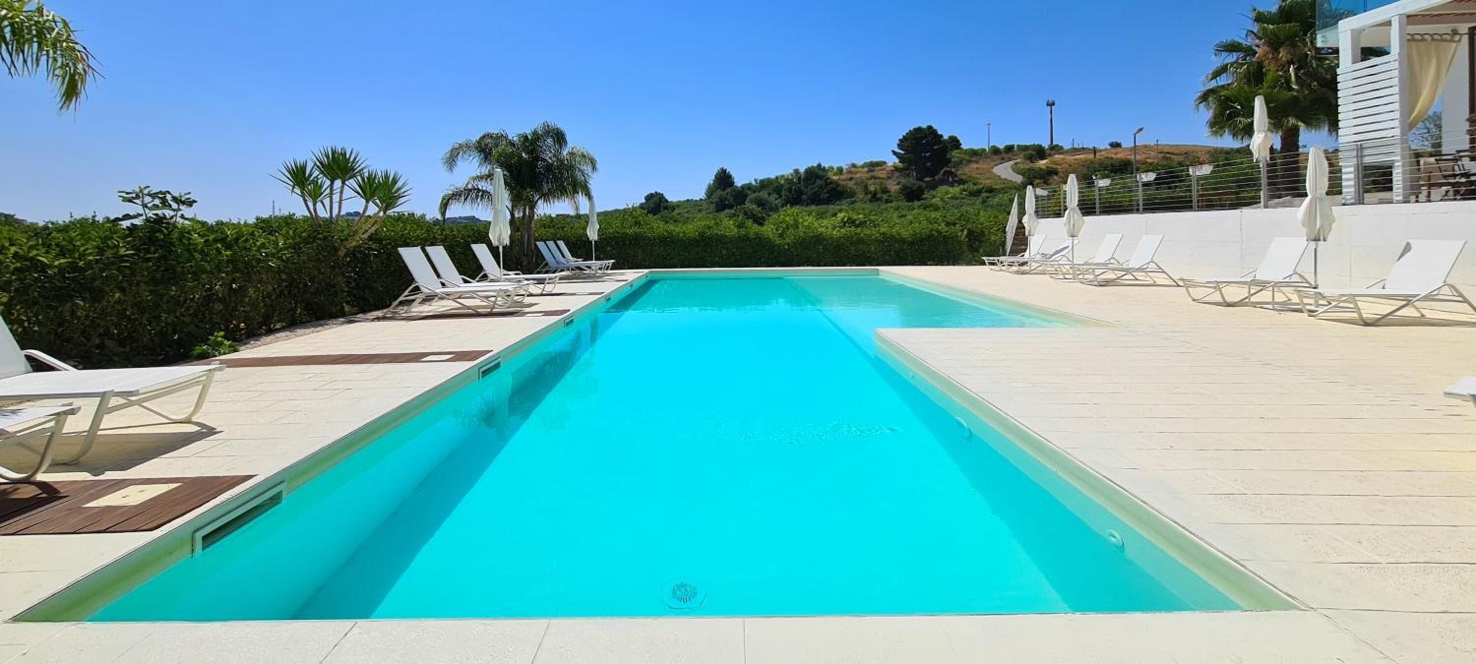 Una piscina rectangular con agua azul cristalina, rodeada de sillas de descanso y sombrillas blancas, con un fondo de vegetación y cielo despejado.