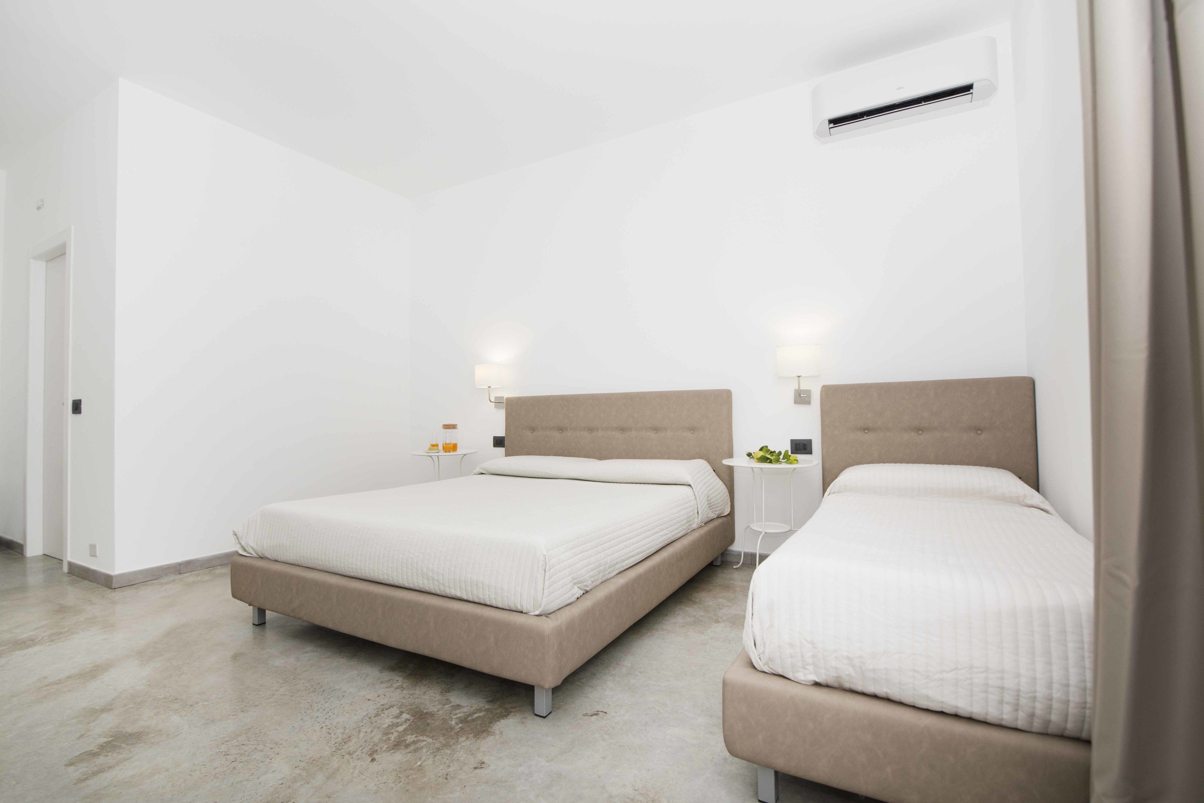 Habitación de hotel moderna con dos camas, una mesita de noche pequeña, aire acondicionado de pared y una puerta a la izquierda.