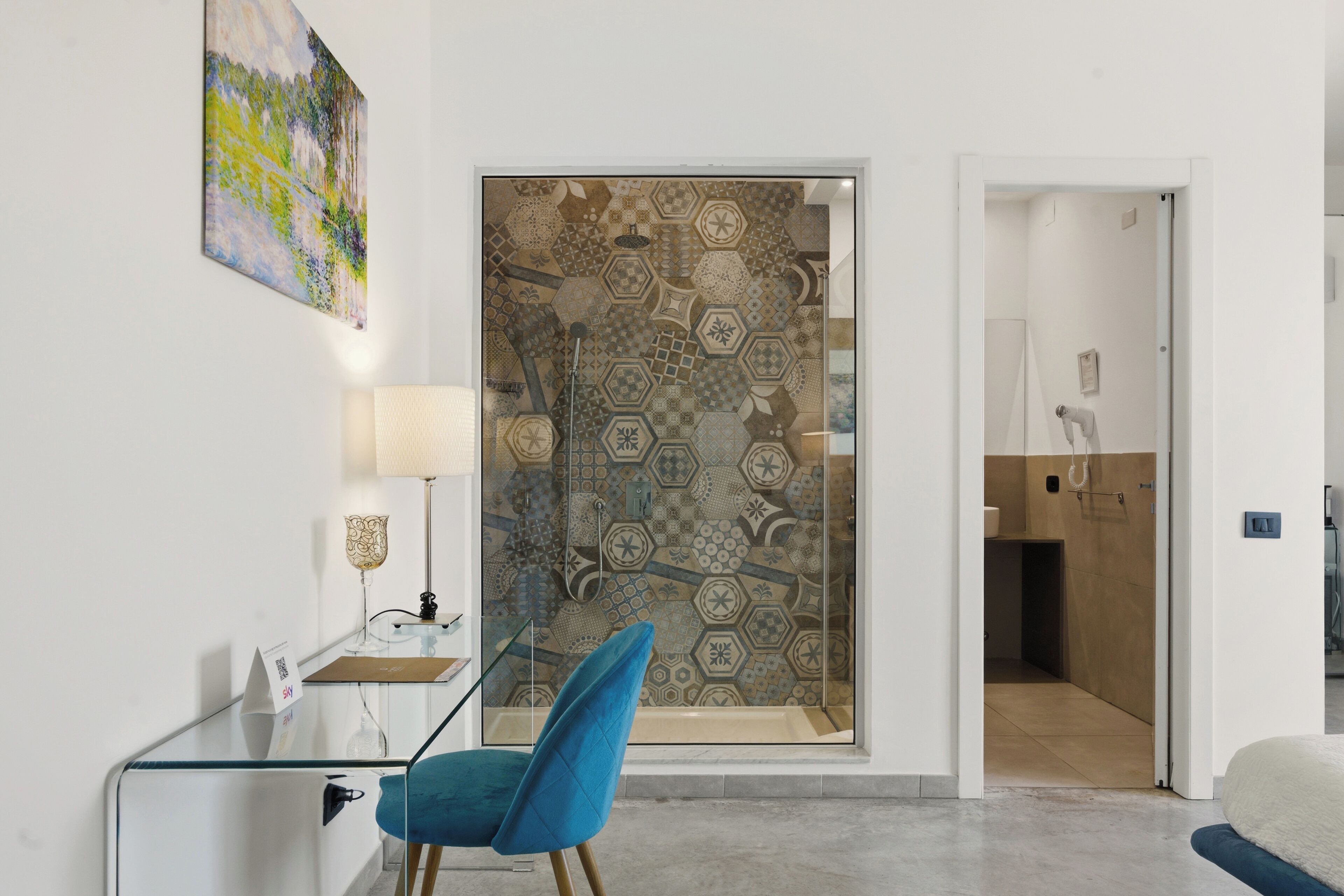 A modern hotel room with a glass partition, a blue chair, a desk with a lamp, and a painting on the wall.