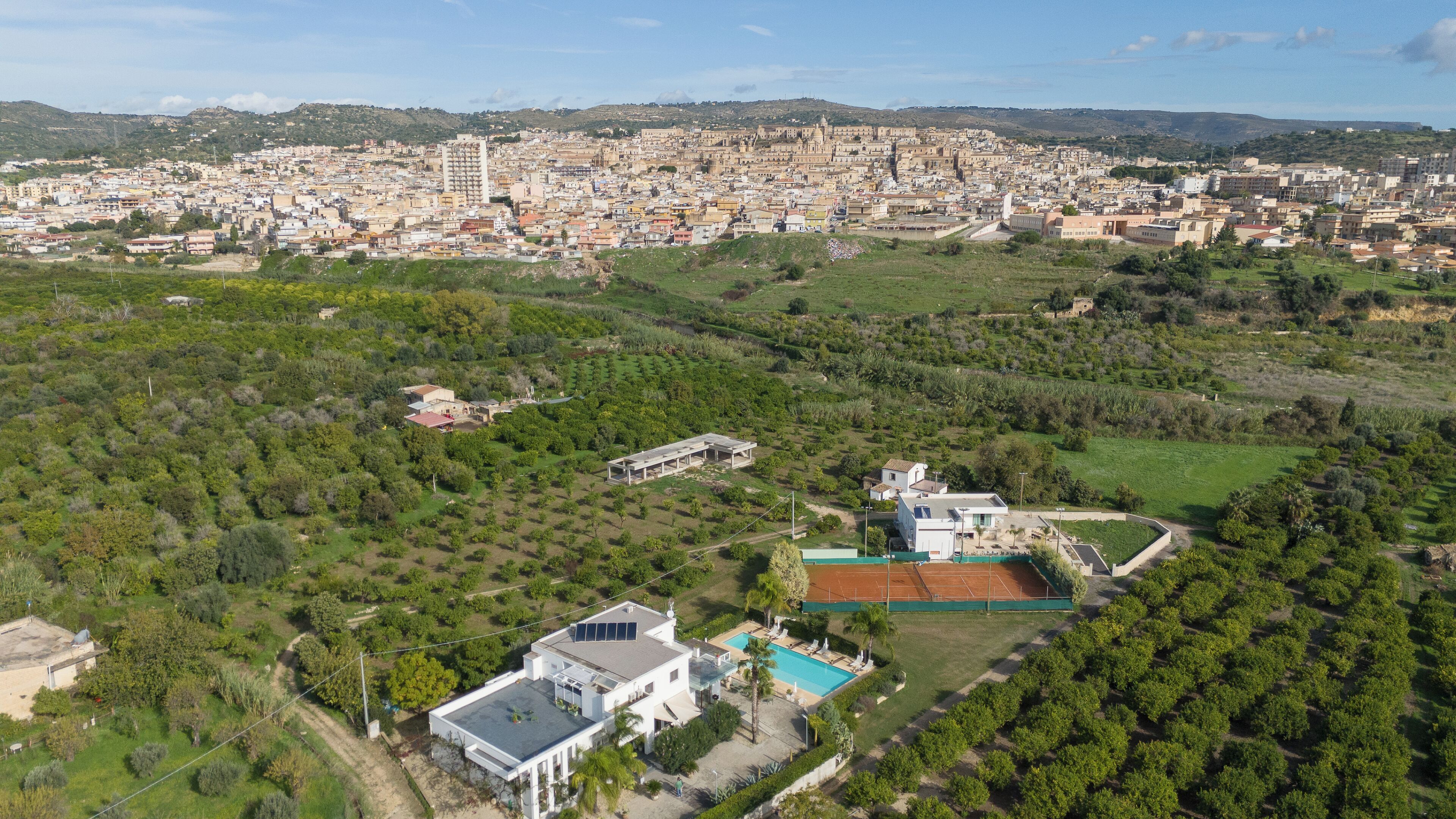Vista aérea de una ciudad que combina zonas urbanas y rurales, con edificios residenciales, una cancha de tenis y espacios verdes.