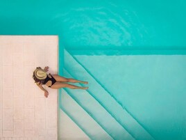 Una persona con traje de baño negro y sombrero de paja parada al borde de una piscina con agua azul cristalina.