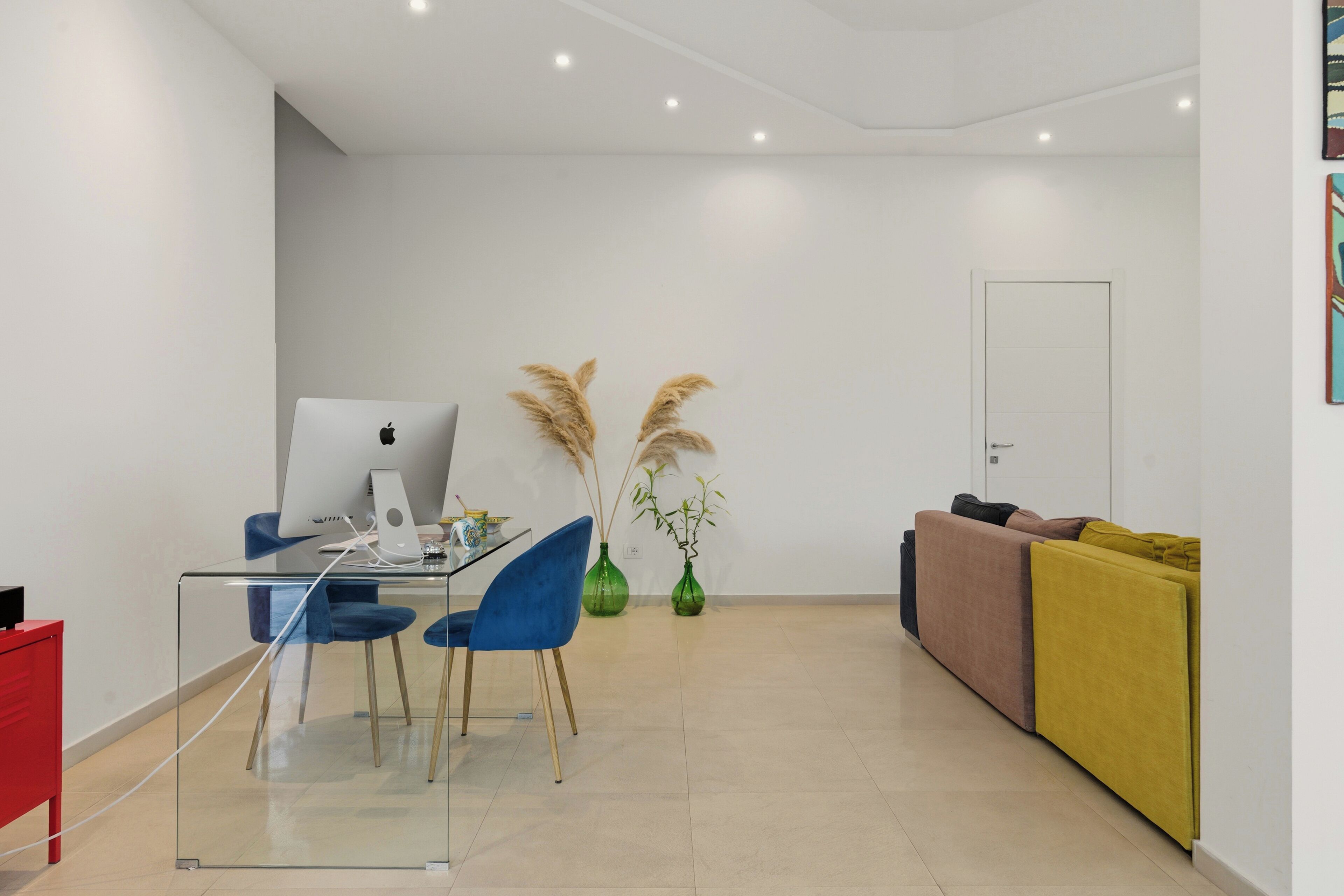 Un espacio de estar moderno con una mesa de comedor de vidrio, sillas azules, una computadora portátil y un sofá con cojines. Hay dos jarrones verdes con plumas de pampa, y una puerta blanca al fondo.