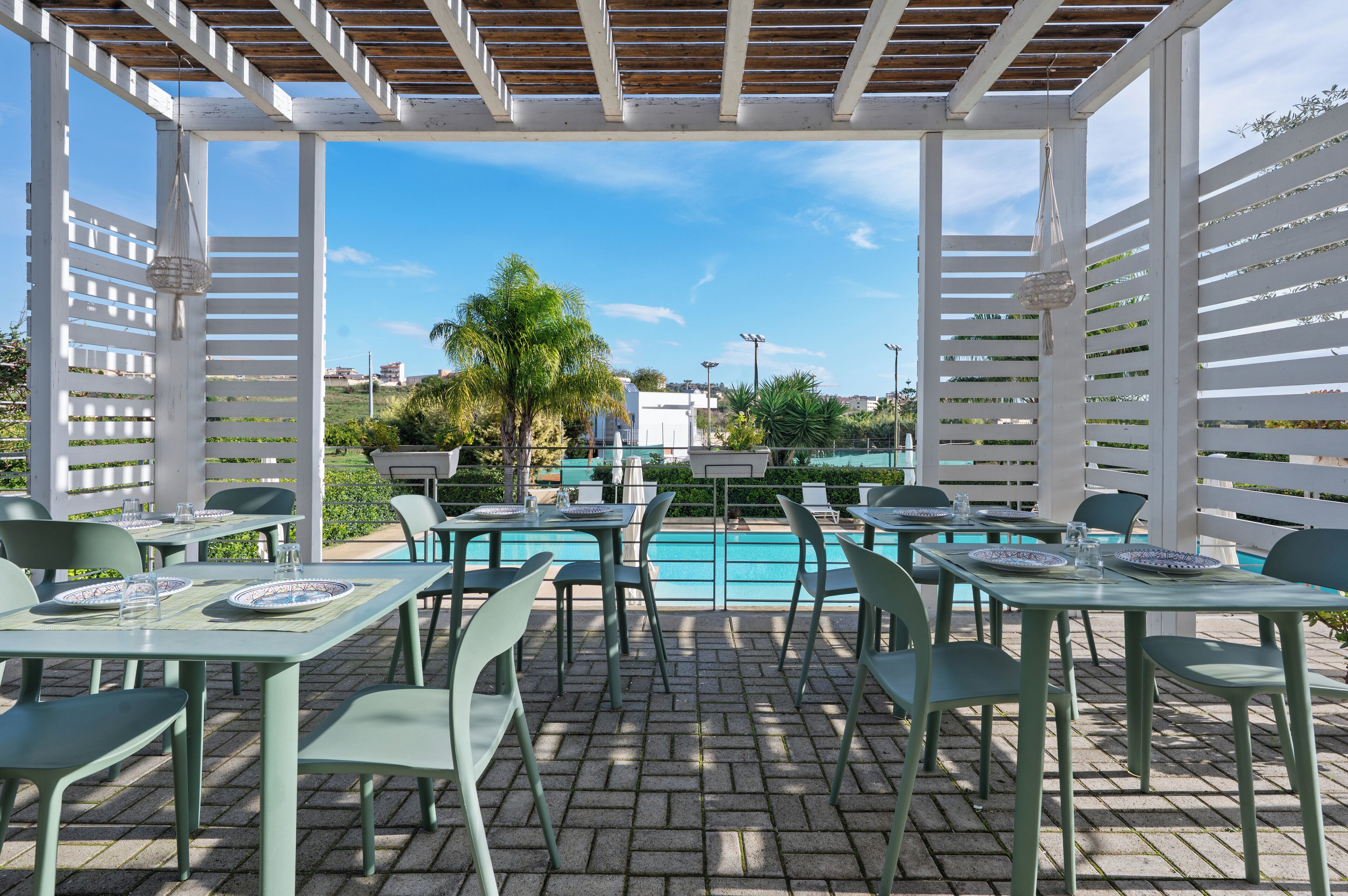 Un espace extérieur abrité, aménagé pour les repas, avec des tables et des chaises, une piscine en arrière-plan et un ciel dégagé.