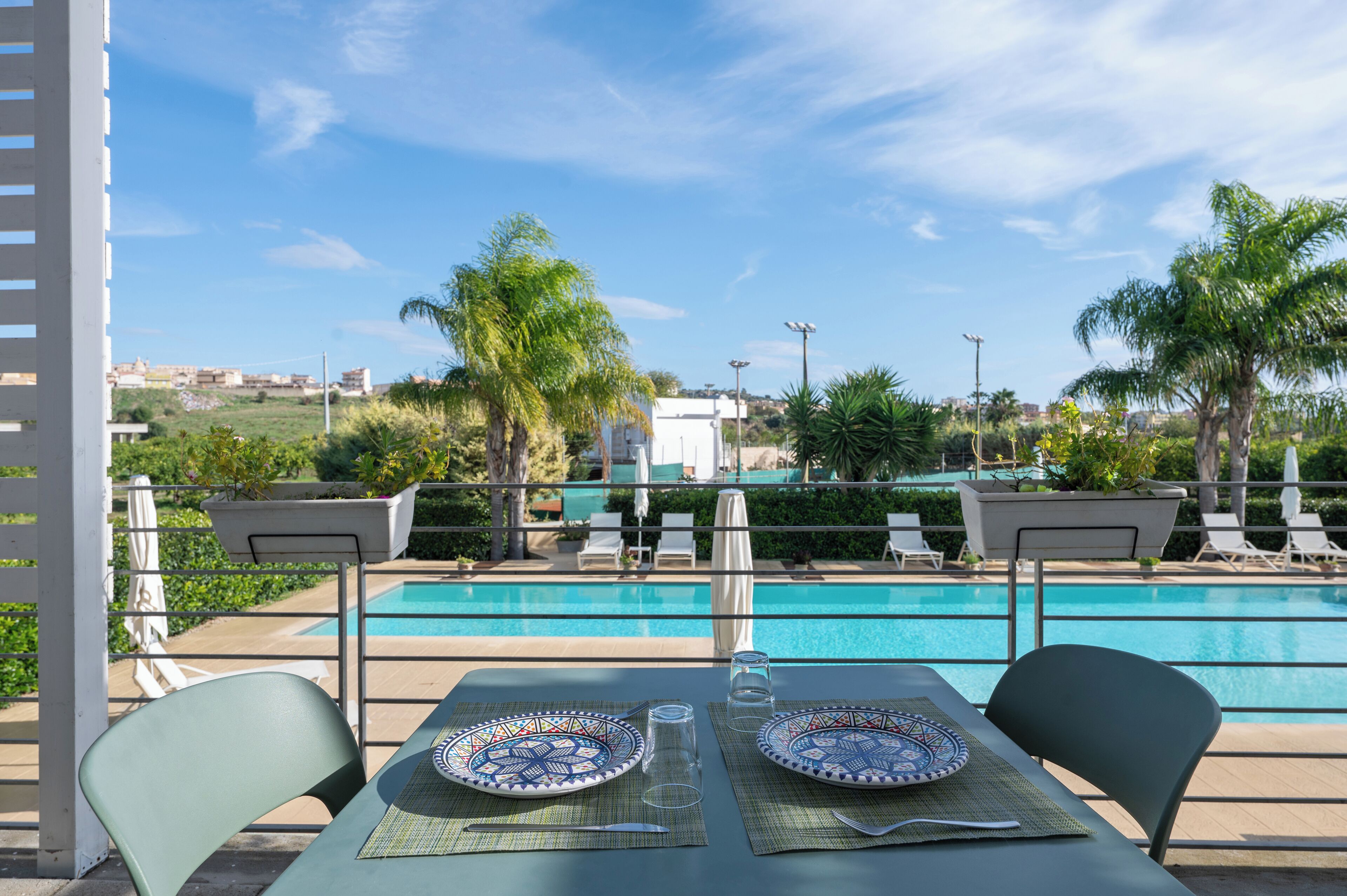 Un comedor con una mesa dispuesta para dos, con vistas a una piscina y sillas de descanso, rodeada de palmeras.