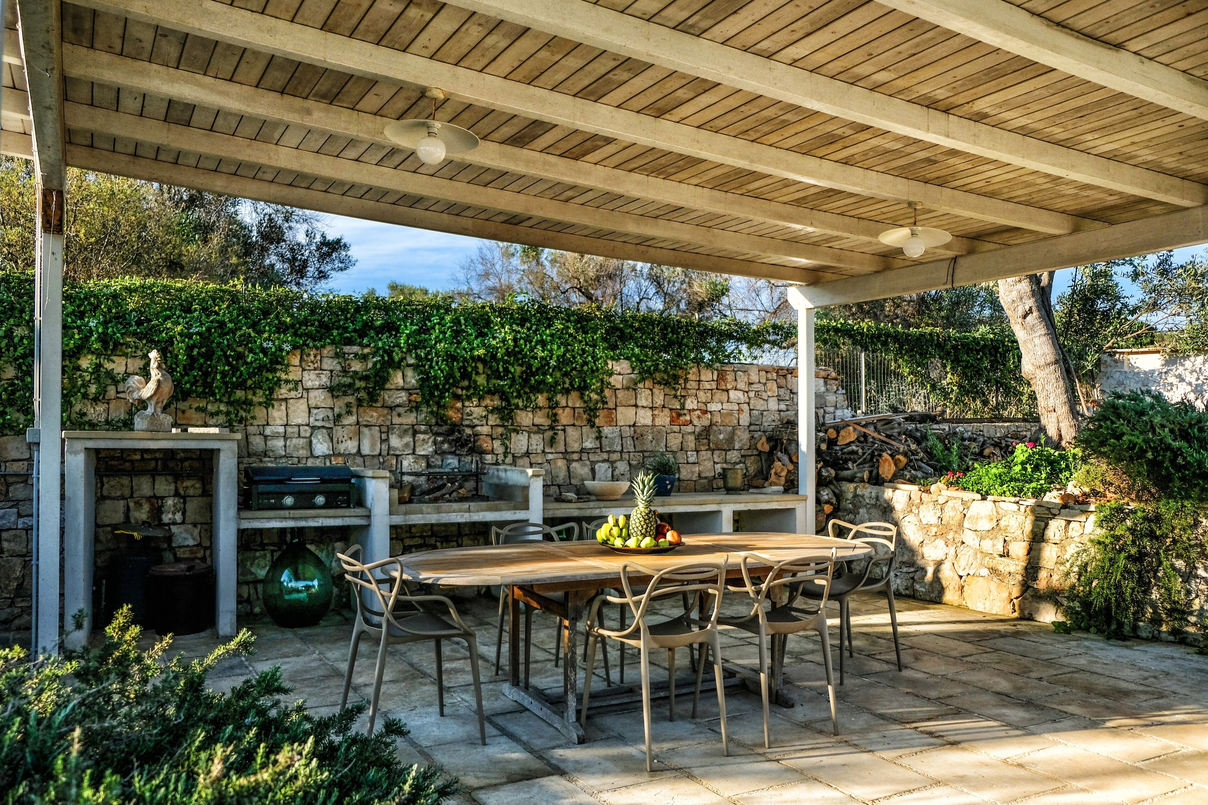 An outdoor patio with a stone wall, a wooden table, and chairs.