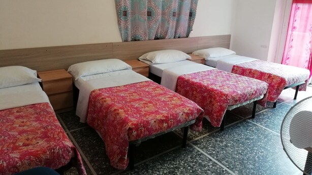 Une chambre d’hôtel avec quatre lits simples, une tête de lit en bois, un tableau au mur, des rideaux à motifs et un sol carrelé.