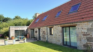 Une maison en pierre avec un toit en pente, de grandes fenêtres et une cour couverte d’herbe.