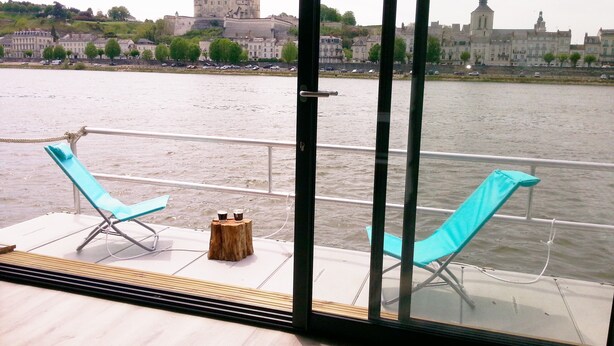 A balcony with two blue lounge chairs and a small wooden stool, overlooking a river with a historic castle and church in the background.