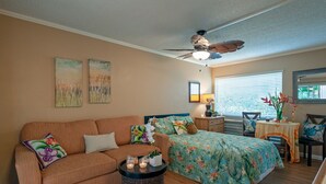 A cozy bedroom with a bed, a sofa, a small table with candles, and a ceiling fan. - Cheerful & Affordable Studio Apartment - Walking Distance to Beaches (Princeville)