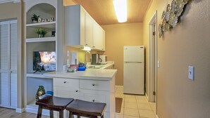 A compact kitchen with white cabinets, a built-in microwave, and a refrigerator. There is a small dining area with two stools and a TV displaying a scenic view. - Cheerful & Affordable Studio Apartment - Walking Distance to Beaches (Princeville)