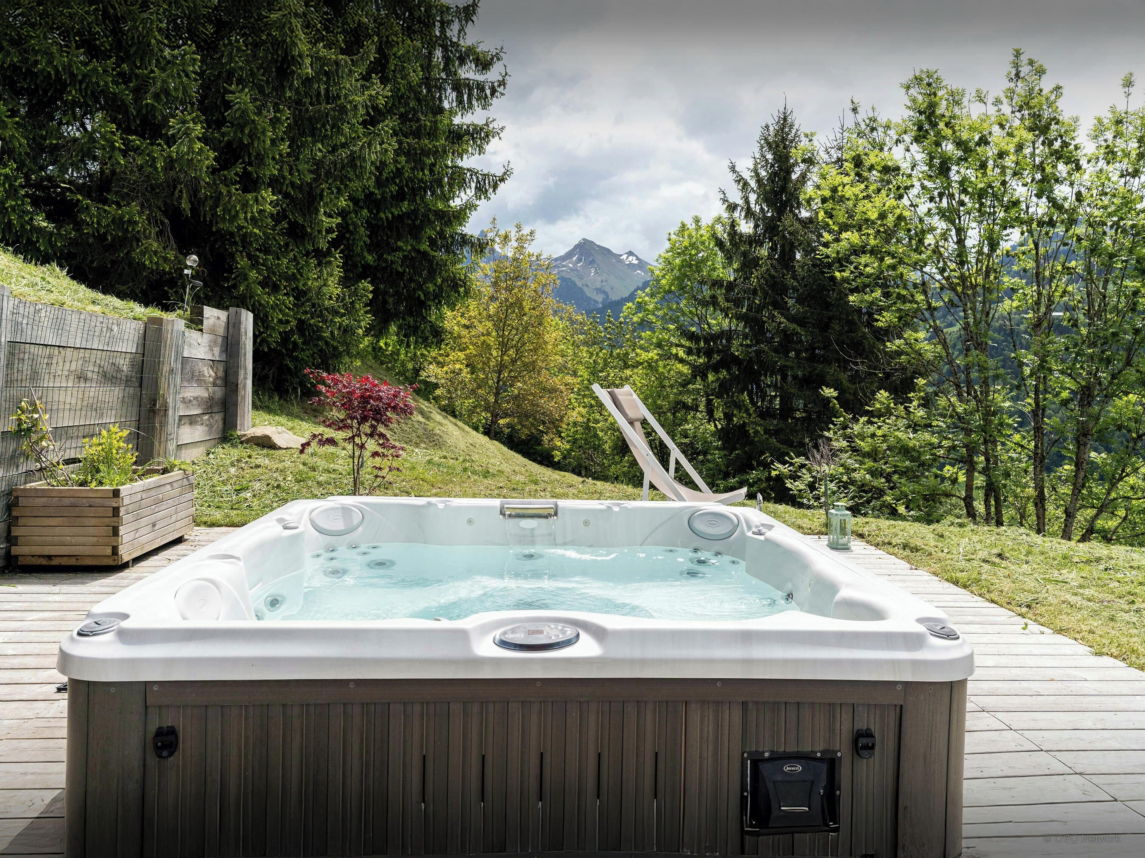 A hot tub in an outdoor setting with a wooden deck, surrounded by greenery and a mountain view.