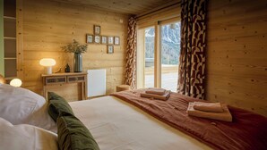 A wooden cabin bedroom with a bed, pillows, a bedside table with a lamp, a vase with flowers, and framed botanical prints on the wall.