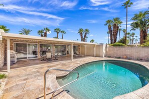 A swimming pool with a stone deck, a covered patio area with chairs and a table, and palm trees in the background. - El Paseo, 'The Rodeo Drive of Beverly Hills' In Palm Desert. Free  Wi FI (Palm Desert)