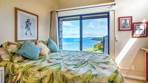 A bedroom with a bed, a large window with a view of the sea, and framed pictures on the wall.