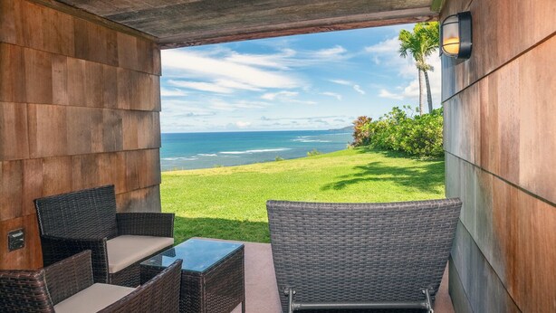 A covered outdoor seating area with wicker furniture and a glass table, overlooking a grassy landscape and the ocean.