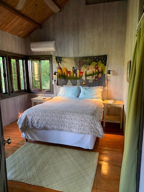 A bedroom with a bed, a nightstand, a wall-mounted air conditioner, a window with blinds, and a painting above the bed.
