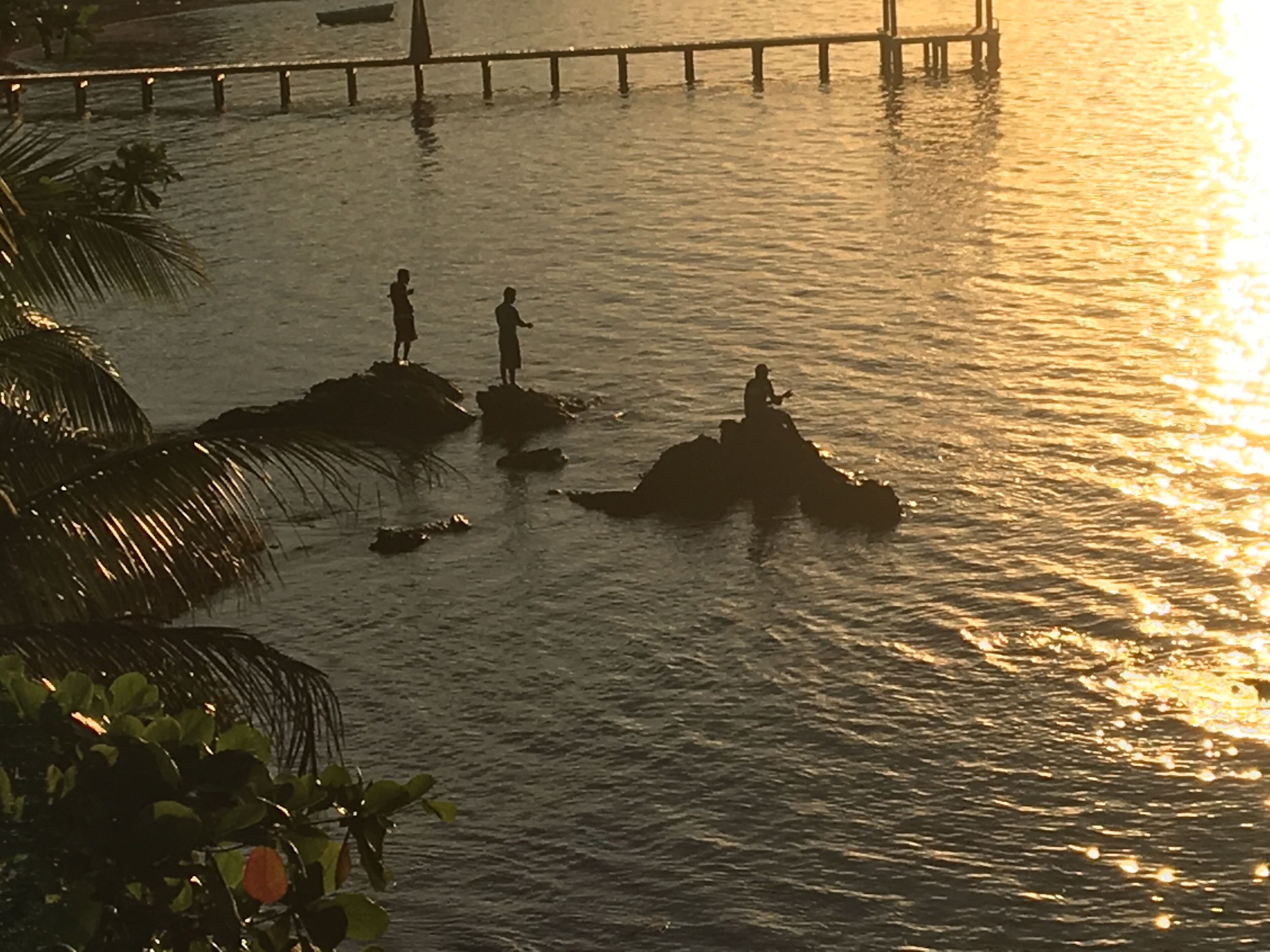 Een zonsondergang boven kalm water, met drie personen op rotsen.