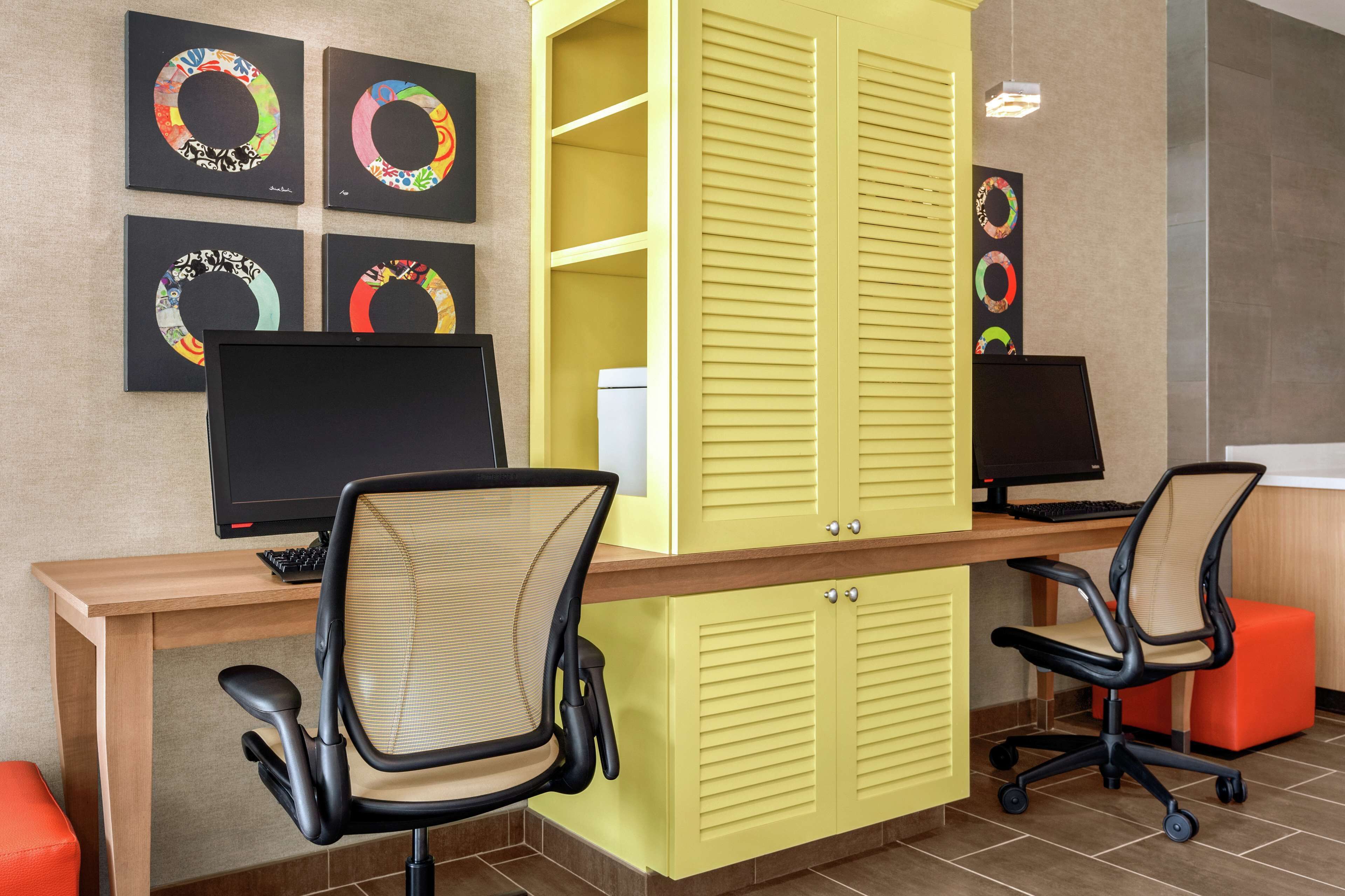 A modern office space with a wooden desk, two black office chairs, a yellow filing cabinet, and four framed circular artworks on the wall.