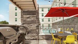 An outdoor patio with a stone wall, a barbecue grill, yellow chairs, and a red umbrella. - Home2 Suites by Hilton Sarasota - Bradenton Airport, FL (Sarasota)