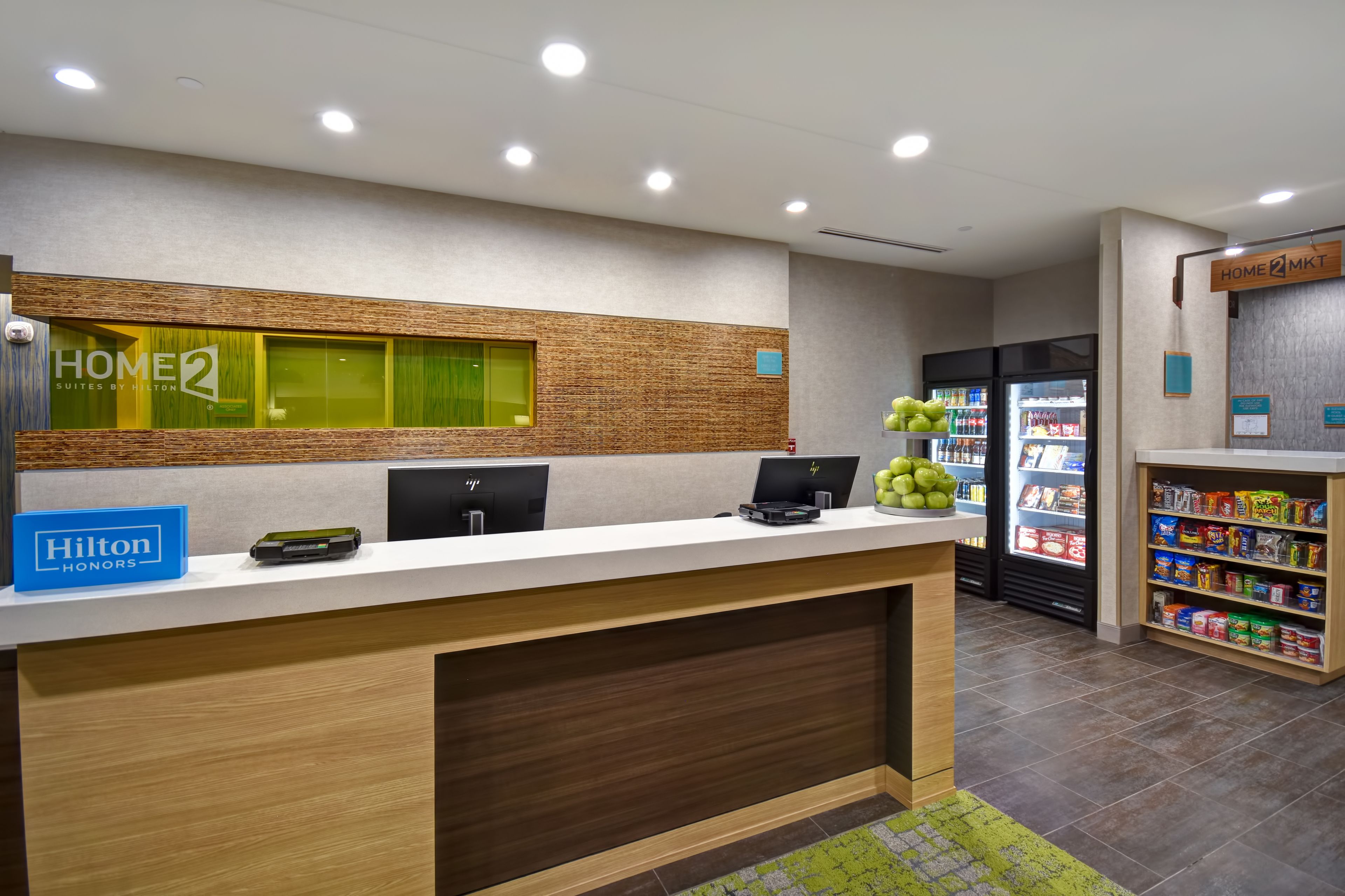Zona de recepción de hotel con un mostrador de madera, un ventanal con marco verde y un refrigerador lleno de bebidas.