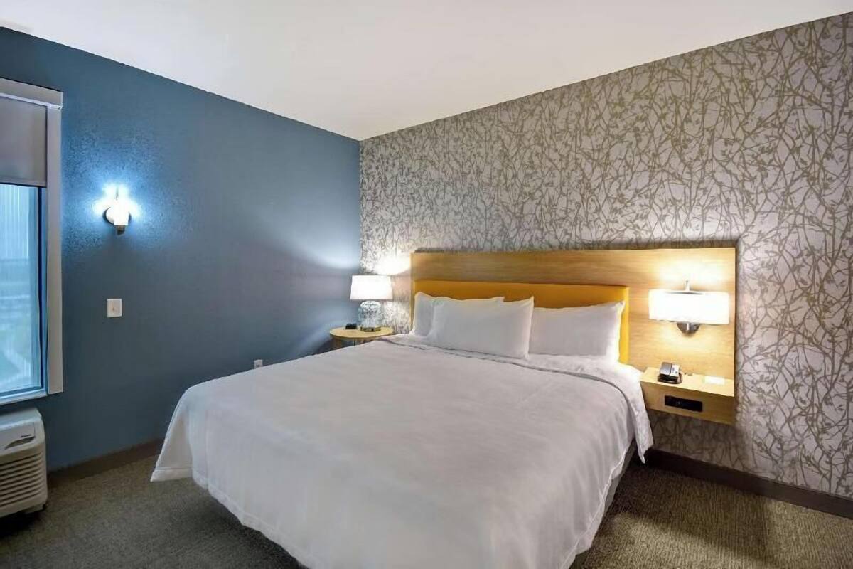 Una habitación de hotel con una cama grande, dos lámparas de noche, una mesita de noche, una ventana y un papel tapiz estampado.