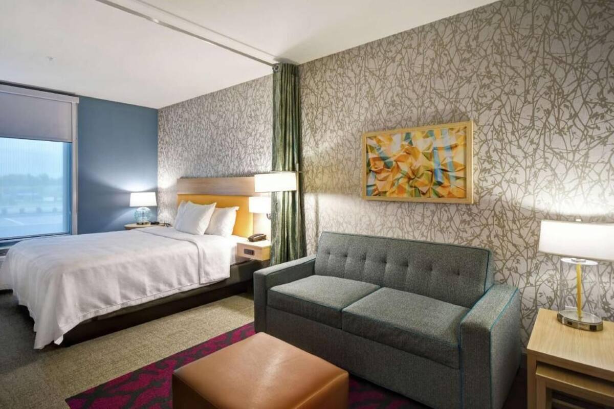 Una habitación de hotel con cama, sofá, una mesita, lámpara y un cuadro en la pared.