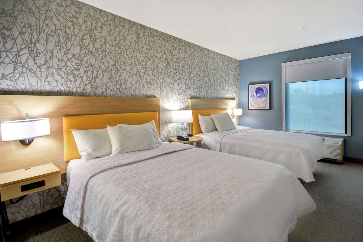 Una habitación de hotel con dos camas, una mesita de noche con lámpara, un televisor montado en la pared y un ventanal grande.