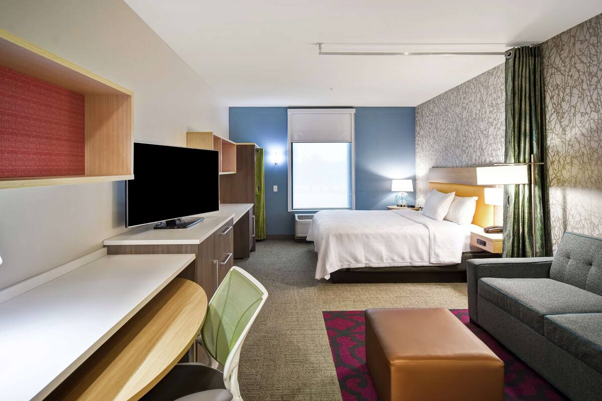 Habitación de hotel con cama, escritorio con silla, sofá, televisor y ventana con cortinas.