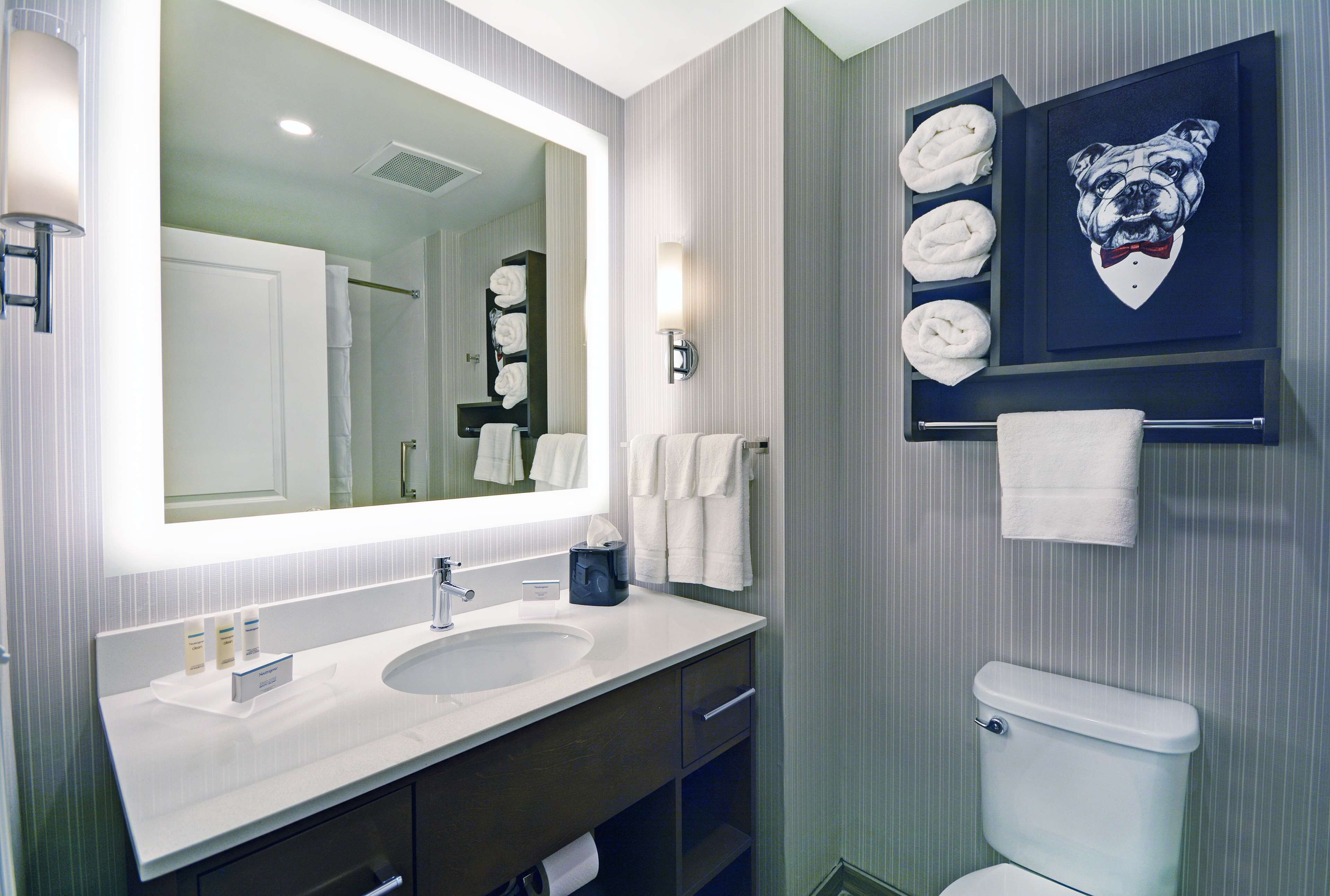 A bathroom with a large mirror, white sink, and a toilet. There are towels and a framed picture on the wall.