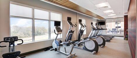 A gym with stationary bikes, treadmills, and a large window offering a view of trees and a road.
