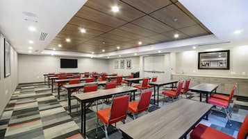 A conference room with multiple tables and chairs, a flat-screen TV, and a kitchen area.