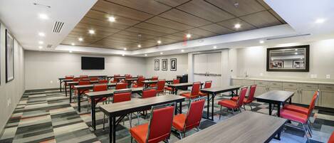 A conference room with multiple tables and chairs, a flat-screen TV, and a kitchen area.