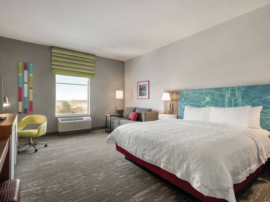 Une chambre d’hôtel avec un grand lit, un bureau, une chaise, une fenêtre avec des stores, un mur orné d’un tableau et une table de chevet avec une lampe.